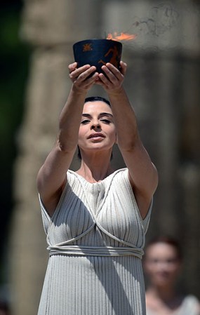 La actriz Ino Menegaki, vestida como una sacerdotisa, estuvo de pie frente al Templo de Hera, de 2.600 años de antigüedad, y después de una invocación a Apolo, el antiguo Dios Sol de los griegos, utilizó un espejo para concentrar los rayos de astro y encender una antorcha.