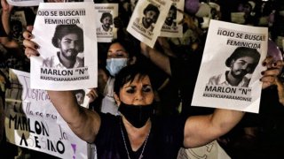 Una mujer, rodeada de más mujeres, alza sus brazos para mostrar dos carteles de un presunto femenicida