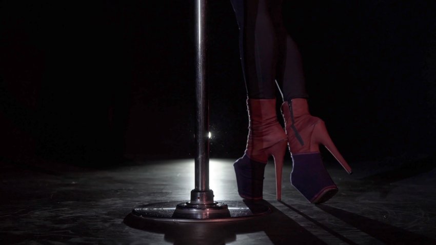 Pole dance. Young beautiful caucasian brunette woman dancer in studio at black background. Girl dancing striptease. Sexy female in shoes on high heels strips, pants and crop top rotates and twirls.
